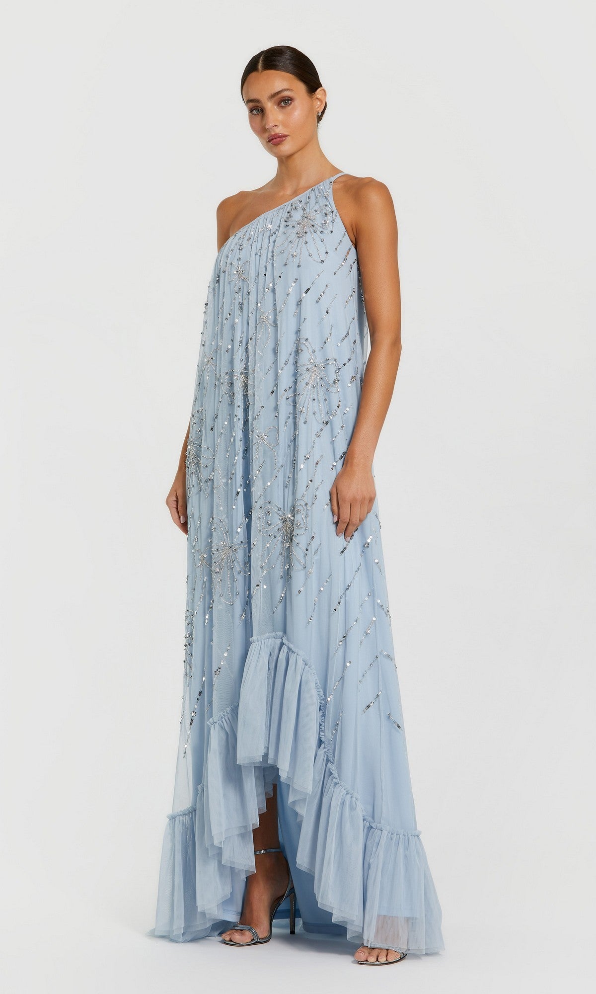 A woman wears the Mac Duggal 9344 Long Formal Dress in powder blue, a one-shoulder gown with sequin embellishments, sheer fabric, ruffled hem, and high-heeled sandals.
